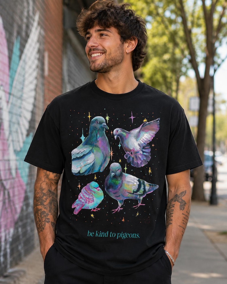 Może przedstawiać: Czarny t-shirt z kr&oacute;tkim rękawem z nadrukiem graficznym przedstawiającym cztery gołębie w opalizujących odcieniach fioletu, zieleni i błękitu, otoczone małymi kolorowymi gwiazdkami. Napis "be kind to pigeons." jest nadrukowany w kolorze morskim.