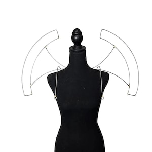 Può includere: Un accessorio con struttura in filo d'argento, forse per costume o esposizione, è esposto su un manichino nero. La struttura in filo ha una forma curva, simile ad ali, con fili sottili che formano la struttura. Il manichino è su sfondo bianco.