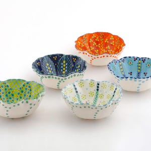 May include: Five decorative ceramic bowls with scalloped edges. Each bowl features a unique color scheme: green, blue, orange, and white, with hand-painted floral patterns and dotted rims. These bowls are ideal for serving snacks or as decorative accents.