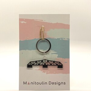 May include: A black acrylic keychain featuring a silhouette of a bridge. The keychain is attached to a silver key ring and has a brown twine string. The keychain is displayed on a card with a pink, blue, and white watercolor background. The card also has the text "Manitoulin Designs" printed on it.