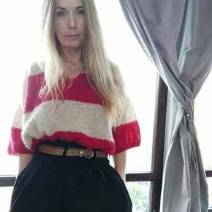 Op de afbeelding: Een vrouw draagt een roze en wit gestreepte trui met een bruine leren riem. Ze staat voor een raam met een grijs gordijn.
