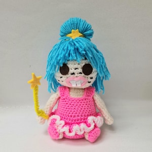 May include: A handmade crochet doll with blue yarn hair, a yellow star hair accessory, and a yellow star wand. The doll wears a pink dress with white trim and has large brown eyes and a pink mouth.