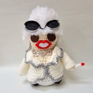 May include: A handmade crochet doll resembling a glamorous figure, wearing sunglasses, a white jacket with silver trim, and a sparkling necklace. The doll has a red lipstick and is holding a cigarette.