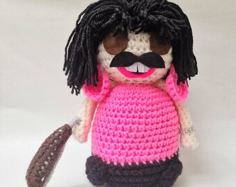 May include: Crocheted doll with black hair, a black mustache, and a pink dress. The doll has a silver bracelet and is holding a brown and yellow object. The doll's mouth is open, revealing two white teeth.