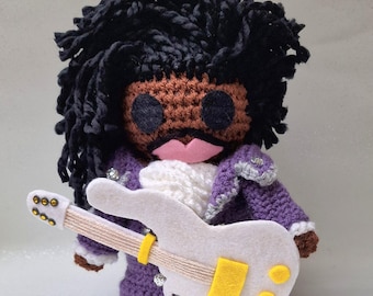May include: A handmade crochet doll of a musician with black yarn hair, brown skin, and a pink mouth. The doll wears a purple jacket with silver accents and holds a white guitar with yellow details. The doll's eyes are black.