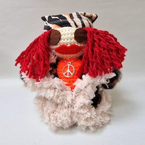 May include: A handmade doll with a crocheted face, red yarn hair, and a large red mouth. The doll wears a necklace with a peace sign pendant and a fluffy white and brown coat. The doll's head is topped with a patterned fabric.