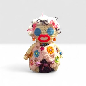 Peut inclure: Poupée au crochet faite à la main avec un design fantaisiste. La poupée présente des yeux bleus, des lèvres rouges et une perruque bouclée blanche avec des accents roses. Elle est ornée de fleurs colorées, de bijoux et d'un nœud violet.