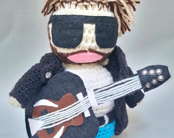 May include: A crocheted plush toy of a person wearing sunglasses and a black leather jacket, holding a black and brown guitar. The toy has brown and white hair and a pink mouth.