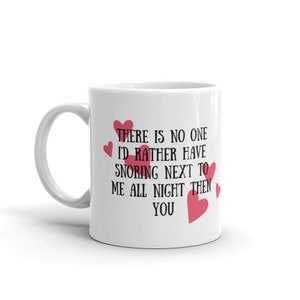 Puede incluir: Taza de café de cerámica blanca con un diseño en negro y rojo. La taza tiene un asa y dice "THERE IS NO ONE I'D RATHER HAVE SNORING NEXT TO ME ALL NIGHT THEN YOU" con corazones rojos.