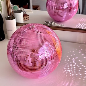 May include: Two pink disco balls with mirrored surfaces. The balls are round and reflect light, creating a sparkling effect. One ball is in the foreground, and another is reflected in a mirror. The balls are on a white surface.