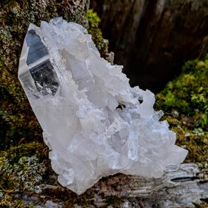 May include: A large, clear quartz crystal cluster with a prominent point. The crystal is surrounded by smaller crystals and has a rough, natural texture.