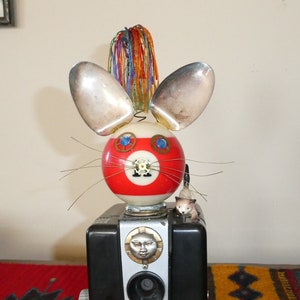 May include: A whimsical sculpture of a rabbit-like creature made from repurposed objects. The head is a red and white striped billiard ball with spoon ears and wire whiskers. The body is a black camera with the text "Brownie Hawkeye Camera Flash Model".