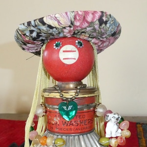 May include: A whimsical doll made from recycled materials. The doll has a red head with green eyes, a floral fabric hat, and a silver metal skirt. The doll is wearing a necklace with a green heart pendant and a beaded necklace with colorful beads. The doll is standing on a red and black rug.