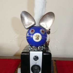 May include: A blue and white 8-ball with red jewels for eyes is the head of a cat sculpture. The cat has white fur for a tail, silver spoons for ears, and a turquoise and silver necklace. The cat is sitting on a black and silver vintage Brownie Hawkeye Flash camera.