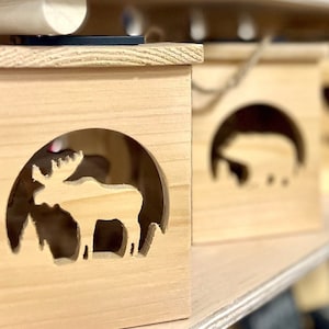 May include: Three wooden boxes with cut-out animal silhouettes. The box on the left features a moose, the middle box features a bear, and the box on the right features a deer.