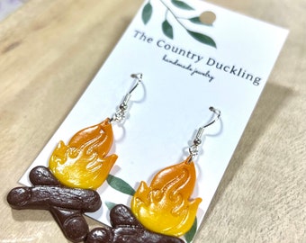 May include: A pair of dangle earrings featuring a campfire design. The earrings are made of polymer clay and have a brown and orange color scheme. The earrings are on a white card with the text "The Country Duckling handmade jewelry".