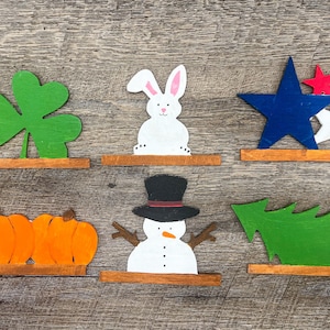 May include: Six wooden cutout decorations for different holidays. A green shamrock, a white bunny, a blue and white star, two orange pumpkins, a white snowman with a black top hat, and a green Christmas tree.