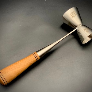 Könnte beinhalten: Ein doppelter Jigger aus Edelstahl mit Holzgriff. Der Jigger hat zwei Messbecher, einer mit "1 1/2 oz" und "3/4 oz" markiert. Der Griff ist hellbraun mit zwei dunkelbraunen Streifen.