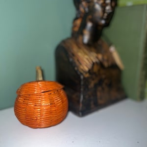 Könnte beinhalten: Ein kleiner orangefarbener, geflochtener Kürbis-Korb mit Deckel. Der Korb steht auf einer weißen Oberfläche vor einer dunkelbraunen Frauenbüste.