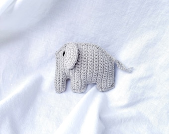 May include: A small, crocheted elephant toy in light gray. The elephant has large ears, a small trunk, and a curled tail. The toy is made of a textured yarn and sits on a white, wrinkled fabric.