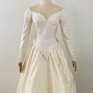 May include: Ivory wedding dress with long sleeves and a full skirt. The bodice is embellished with sequins and beads, and the sleeves have beaded cuffs. The dress features a sweetheart neckline and a bow at the back. The skirt is gathered and falls to the floor.