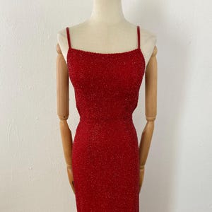 May include: A red beaded evening gown with spaghetti straps. The dress is displayed on a mannequin with wooden arms and legs. The gown has a fitted bodice and a floor-length skirt, perfect for a formal event.