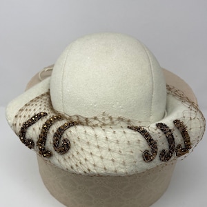 May include: A white felt hat with a brown beaded trim and a sheer veil. The hat is sitting on a beige box.