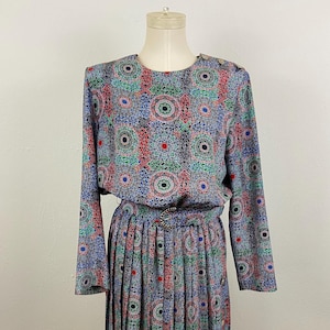 May include: A long-sleeved dress with a pleated skirt. The dress is made of a blue, green, red, and white patterned fabric. The dress has a round neckline and a button closure at the neck.