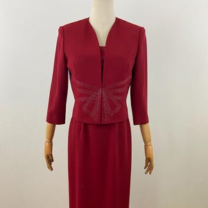 Op de afbeelding: Een formele rode jurkset, bestaande uit een lange rok en een bijpassende jas. De jas heeft driekwart mouwen en een decoratief strass strik ontwerp op de voorkant. De jurk wordt gepresenteerd op een mannequin.