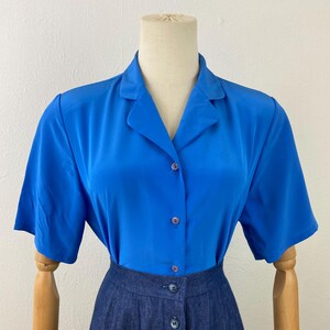 May include: A short-sleeved, cobalt blue blouse with a collared neckline and button-up front is paired with a dark denim skirt. The skirt features a button-down design. The clothing is displayed on a mannequin.