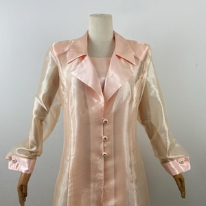 May include: A light pink, sheer, long-sleeved dress with a collared neckline and button-down front. The dress features a satin collar, cuffs, and buttons. The dress is displayed on a mannequin.
