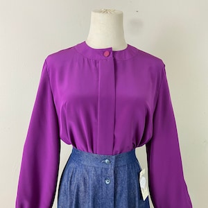 May include: A purple long-sleeve blouse with a button-up front and a round collar. The blouse has a slightly puffed sleeve. The blouse is paired with a blue denim skirt with a button-up front.