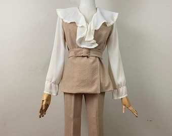 May include: A beige pantsuit with a ruffled white blouse. The pantsuit has a wide leg and a belt. The blouse has long sleeves and a ruffled collar.