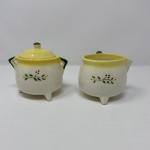 May include: A white ceramic sugar and creamer set with a yellow rim and green accents. The set features a floral design with green leaves, yellow flowers, and red berries.