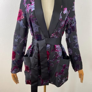May include: A black blazer featuring a floral pattern in shades of magenta and purple. The garment includes a solid black satin shawl collar, a matching black satin tie belt at the waist, and two large patch pockets on the front lower section.