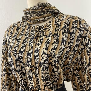 Peut inclure: Un chemisier vintage &agrave; motif peau de serpent noir, beige et cr&egrave;me. Le chemisier a des manches longues, un col montant et des boutons dor&eacute;s. Une &eacute;charpe assortie est drap&eacute;e autour du cou. Le bas du chemisier est associ&eacute; &agrave; une jupe noire.