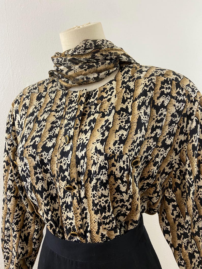 Peut inclure: Un chemisier vintage &agrave; motif peau de serpent noir, beige et cr&egrave;me. Le chemisier a des manches longues, un col montant et des boutons dor&eacute;s. Une &eacute;charpe assortie est drap&eacute;e autour du cou. Le bas du chemisier est associ&eacute; &agrave; une jupe noire.