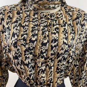 Peut inclure: Un chemisier vintage &agrave; imprim&eacute; animalier noir, beige et cr&egrave;me. Le chemisier a des manches longues, un col montant et des boutons dor&eacute;s. Le v&ecirc;tement est associ&eacute; &agrave; une jupe noire, cr&eacute;ant un look r&eacute;tro classique.
