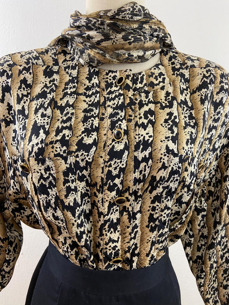 Peut inclure: Un chemisier vintage &agrave; imprim&eacute; animalier noir, beige et cr&egrave;me. Le chemisier a des manches longues, un col montant et des boutons dor&eacute;s. Le v&ecirc;tement est associ&eacute; &agrave; une jupe noire, cr&eacute;ant un look r&eacute;tro classique.