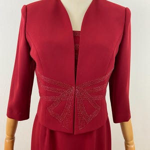 Op de afbeelding: Een rode tweedelige jurkset. De jas heeft driekwartmouwen en een decoratief kralenontwerp over de taille. De jurk eronder is zichtbaar, met een vergelijkbaar kralenpatroon. De set wordt op een mannequin gepresenteerd.