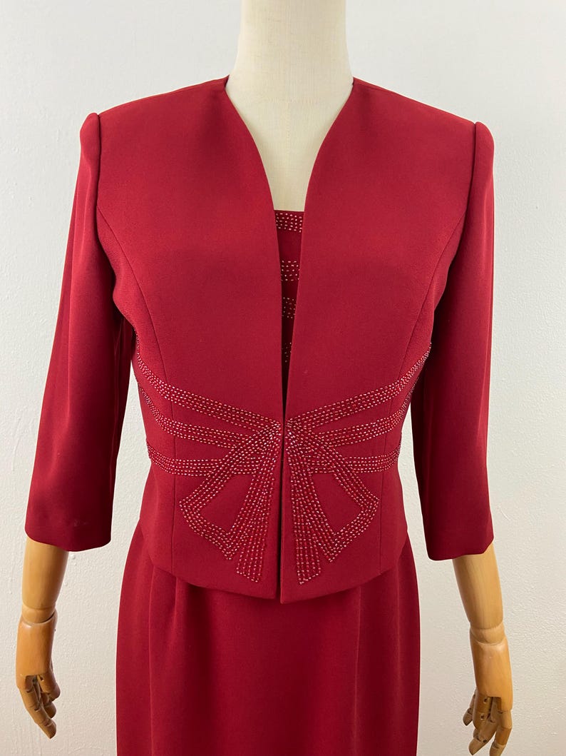 Op de afbeelding: Een rode tweedelige jurkset. De jas heeft driekwartmouwen en een decoratief kralenontwerp over de taille. De jurk eronder is zichtbaar, met een vergelijkbaar kralenpatroon. De set wordt op een mannequin gepresenteerd.