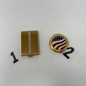 May include: Two gold compact mirrors. One is rectangular with a vertical row of rhinestones. The other is round with a red, white, and blue American flag design.