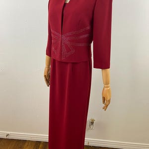 Op de afbeelding: Een rode formele jurkset op een mannequin. De set bevat een lange, rechte rok, een bijpassende jas met driekwartmouwen en een top. De jas heeft een strass-ontwerp aan de voorkant. De jurk wordt gepresenteerd op een houten mannequin.