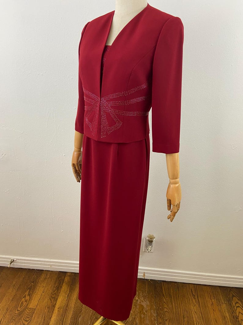 Op de afbeelding: Een rode formele jurkset op een mannequin. De set bevat een lange, rechte rok, een bijpassende jas met driekwartmouwen en een top. De jas heeft een strass-ontwerp aan de voorkant. De jurk wordt gepresenteerd op een houten mannequin.