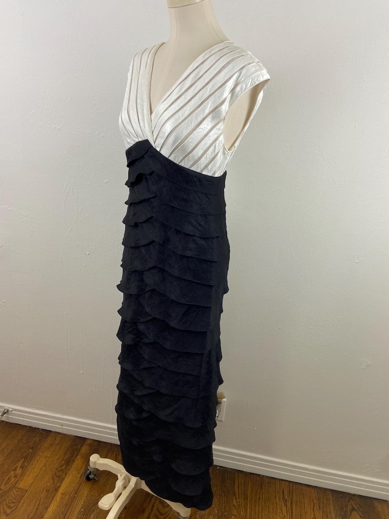 May include: A formal, floor-length dress with a V-neck bodice. The top is white with vertical striped detailing, transitioning to a black, tiered skirt. The dress is displayed on a mannequin.