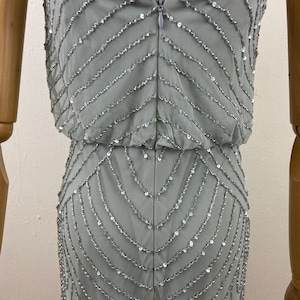 K&ouml;nnte beinhalten: Hellgraues Abendkleid mit eng anliegendem Oberteil und flie&szlig;endem Rock. Das Kleid ist mit Reihen aus silbernen Pailletten verziert, die einen schimmernden Effekt erzeugen. Es hat d&uuml;nne Spaghettitr&auml;ger und einen Rei&szlig;verschluss hinten.
