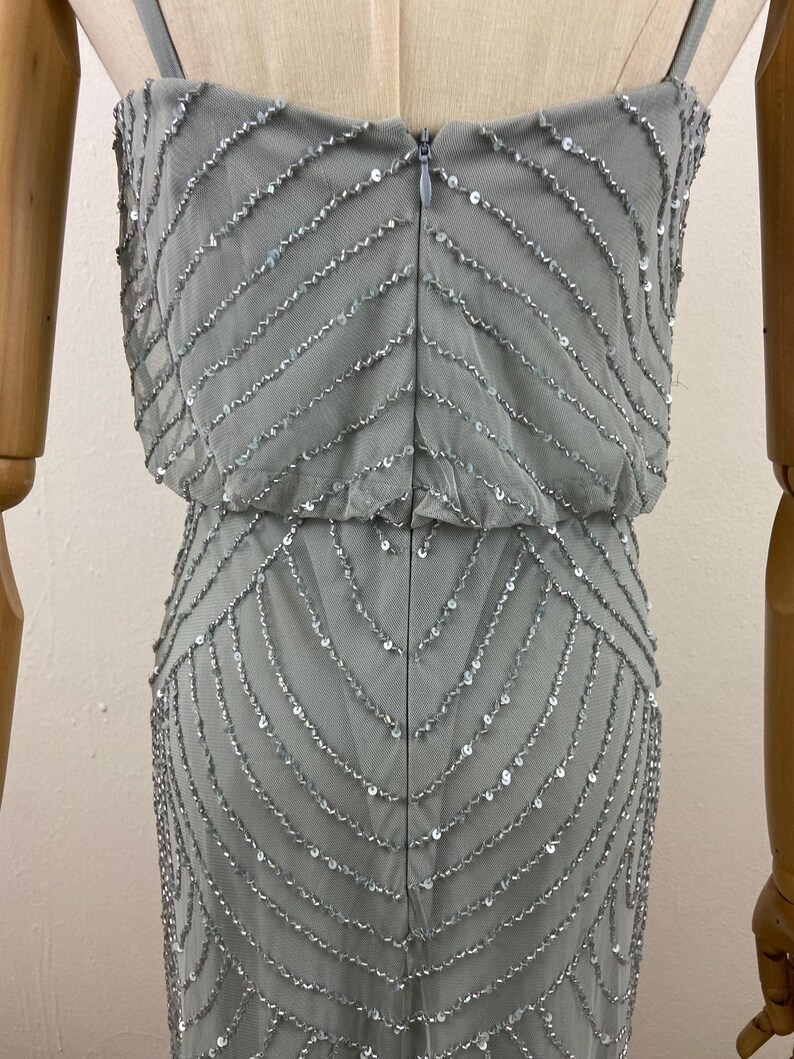 K&ouml;nnte beinhalten: Hellgraues Abendkleid mit eng anliegendem Oberteil und flie&szlig;endem Rock. Das Kleid ist mit Reihen aus silbernen Pailletten verziert, die einen schimmernden Effekt erzeugen. Es hat d&uuml;nne Spaghettitr&auml;ger und einen Rei&szlig;verschluss hinten.