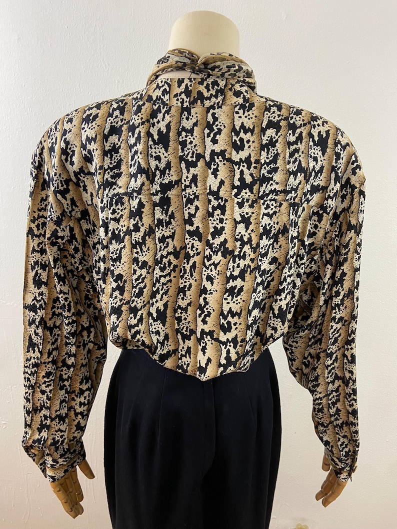 Peut inclure: Un chemisier vintage &agrave; manches longues avec un motif animalier unique dans les tons de marron, noir et cr&egrave;me. Le chemisier pr&eacute;sente un col montant et est associ&eacute; &agrave; un pantalon noir taille haute. Le tissu semble l&eacute;ger et adapt&eacute; &agrave; diverses occasions.