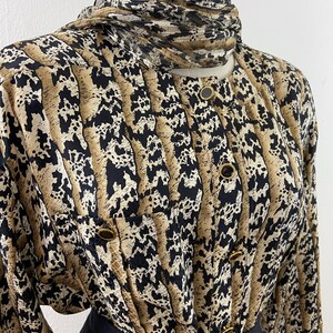 Peut inclure: Un chemisier vintage &agrave; boutons avec un motif unique dans les tons de beige, noir et marron. Le chemisier a un col montant avec une &eacute;charpe assortie et des boutons dor&eacute;s. Les manches sont longues et la partie inf&eacute;rieure est noire.