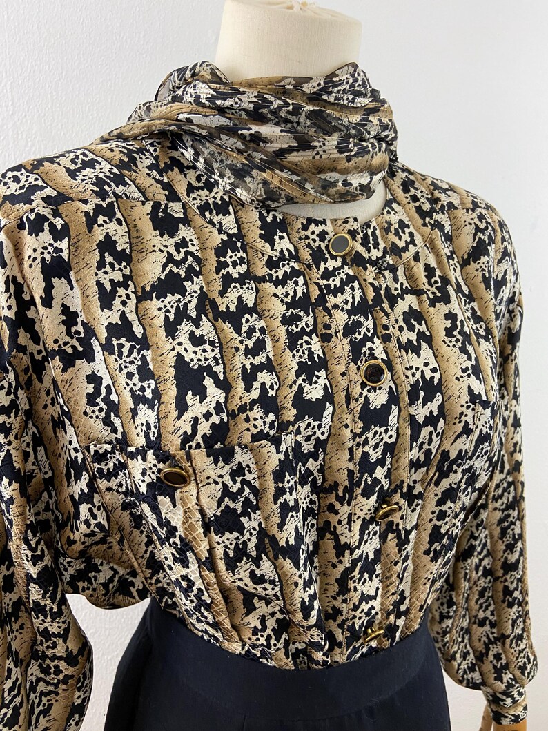 Peut inclure: Un chemisier vintage &agrave; boutons avec un motif unique dans les tons de beige, noir et marron. Le chemisier a un col montant avec une &eacute;charpe assortie et des boutons dor&eacute;s. Les manches sont longues et la partie inf&eacute;rieure est noire.